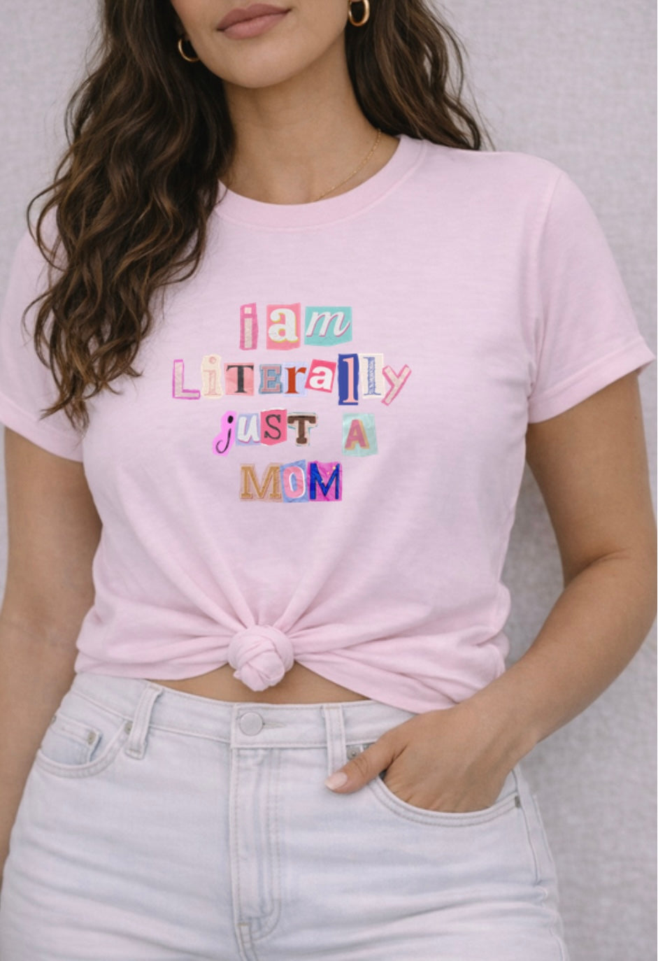 Person wearing a pink t-shirt with colorful text design " I am Literally Just a Mom" against a neutral background