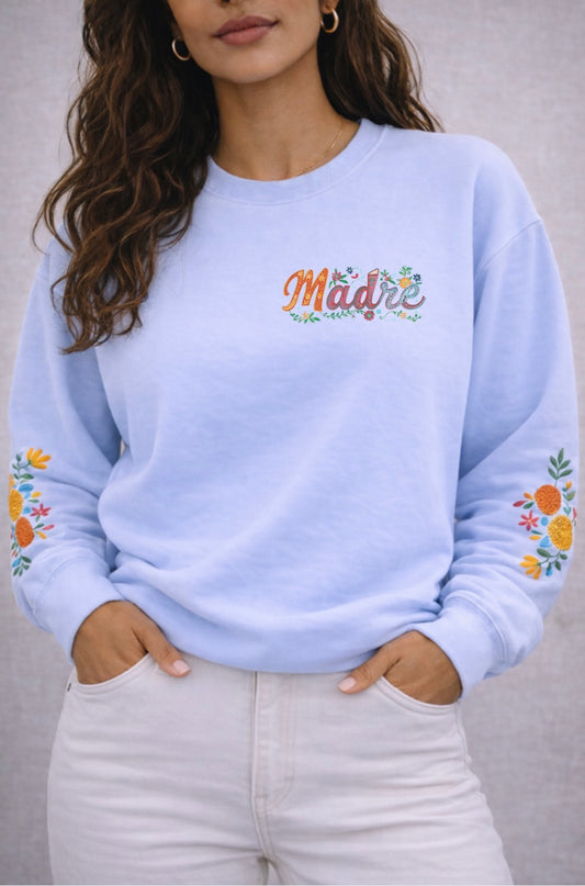 Light blue sweatshirt with floral designs on sleeves and 'Madre' text, worn by a person.