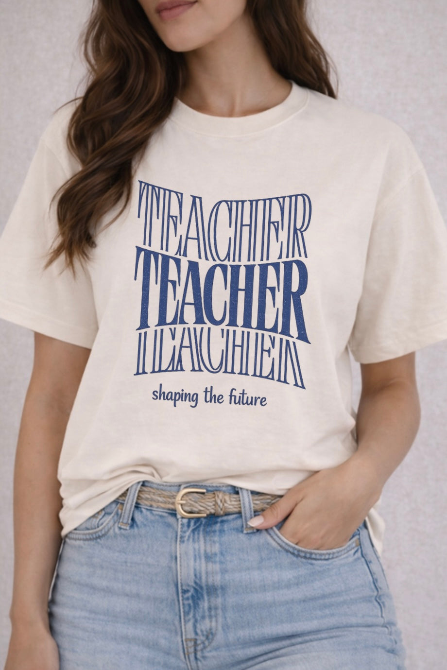 Person wearing a white t-shirt with blue text "Teacher shaping the future" and jeans on a neutral background