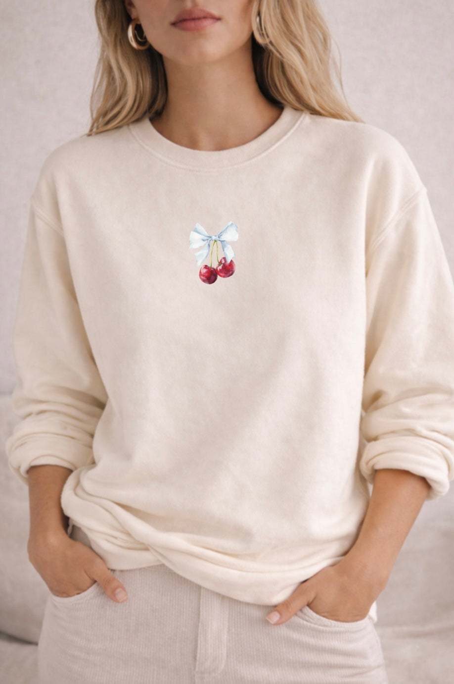 Person wearing a ivory sweatshirt with a cherry with a bow graphic design on a plain background