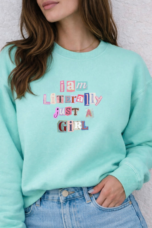 Person wearing a light blue sweatshirt with colorful text design saying "I am literally just a girl", standing against a neutral background.