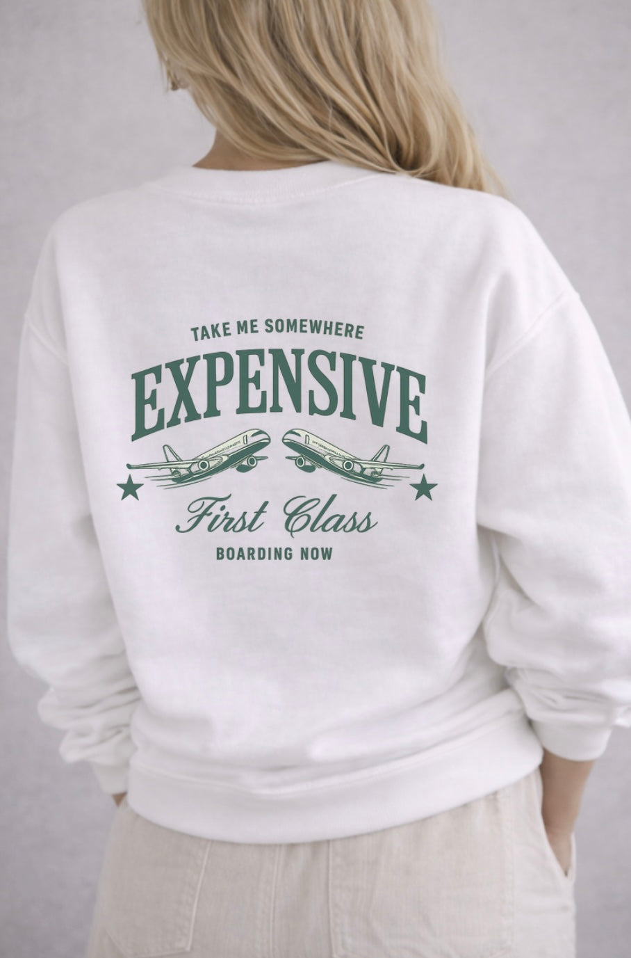White sweatshirt with text "take me somewhere expensive first class boarding now" and airplane graphics on the back, worn by a person.
